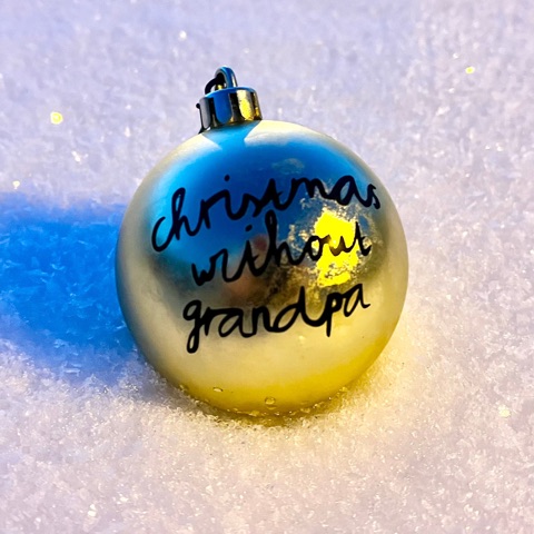 Goldene glu:-Weihnachtsbaumkugel mit schwarzem Schriftzug christmas without grandpa, im Schnee liegend