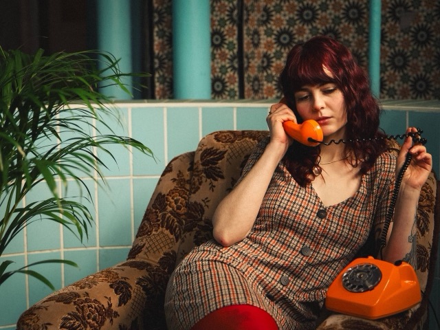Vorschaubild zum Musikvideo smile von glu: – Sängerin Lotti im geblümten Sessel mit orangefarbenem Retro-Telefon